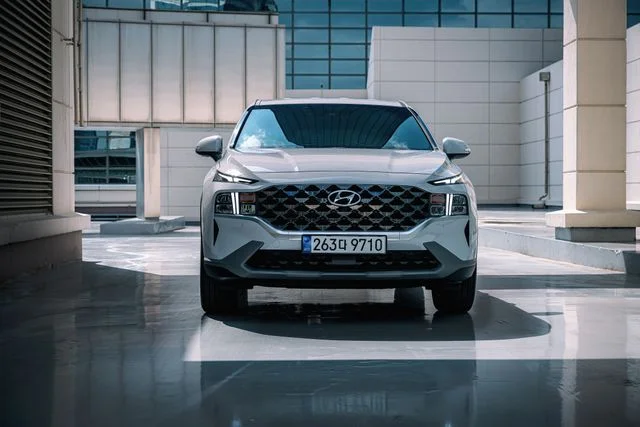Front view of a silver Hyundai SUV parked in a modern urban architectural space, featuring a large black grille, sleek LED headlights, and reflective flooring surrounded by glass and concrete buildings.