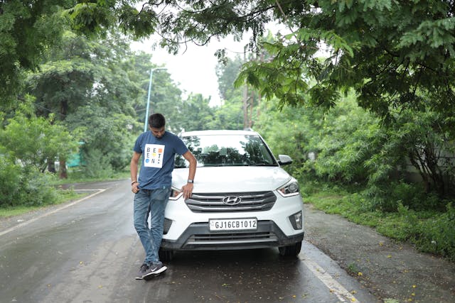 Man standing beside a white Hyundai Creta SUV on a tree-lined road in Gujarat, India